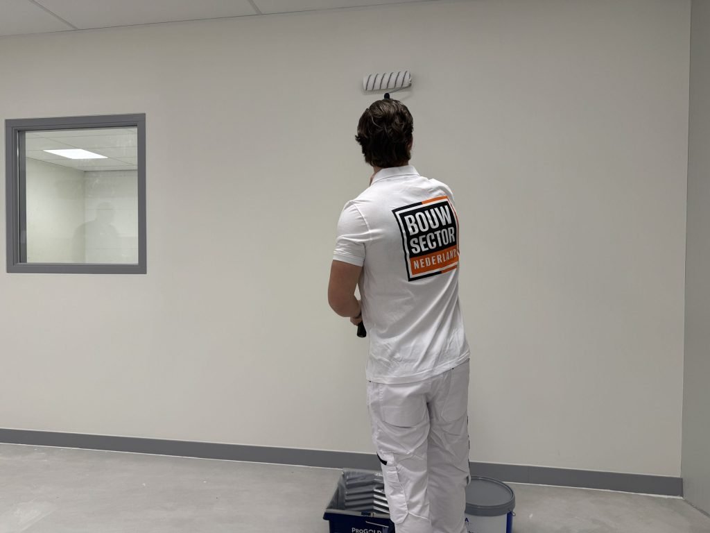 Schilder in witte werkkleding rolt een lichte wand in een lege ruimte, verfemmer en bak op de vloer