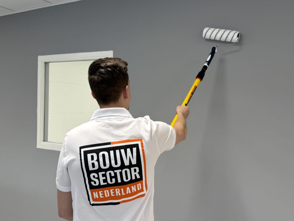 Schilder van Bouwsector Nederland met logo op zijn rug rolt een grijze wand naast een wit kozijn